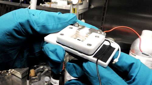 Two hands in blue protective gloves are holding the measuring cell. It is white, about the size of a telephone and connected with cables. 