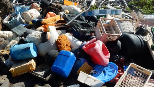 A pile of larger plastic pieces (broken beverage crates, a bobby car, lids from a trash can, baskets, canisters, pipes...) lies on dark volcanic stones.