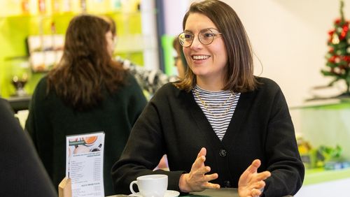 Das Bild zeigt Ann Kathrin Schubert. Sie steht in einem Café an der Uni, im Hintergrund ist die Bar zu erkennen. Sie gestikuliert leicht mit ihren Händen. Sie schaut zu der Interviewerin und lächelt diese an.