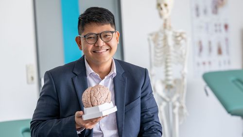 Portrait of Daniel Kristanto. He is wearing glasses and holding a model of a brain. A skeleton stands in the background.