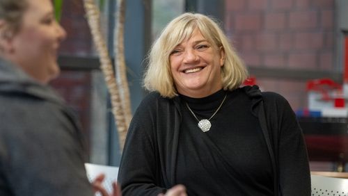 Elke Hoxha lachend während der Interview-Situation. Die Frau mit den schulterlangen blonden Haaren trägt einen schwarzen Rollkragen-Pullover, eine schwarze Strickjacke und eine auffällige, silberne Kette.